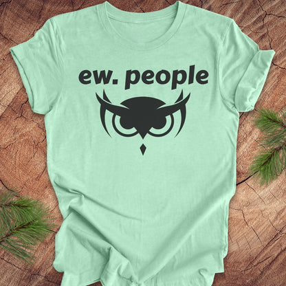 A mint green short-sleeve t-shirt with a black graphic print that reads 'ew. people' above an owl silhouette, laid flat on a rustic wood background with pine branches in the corners.