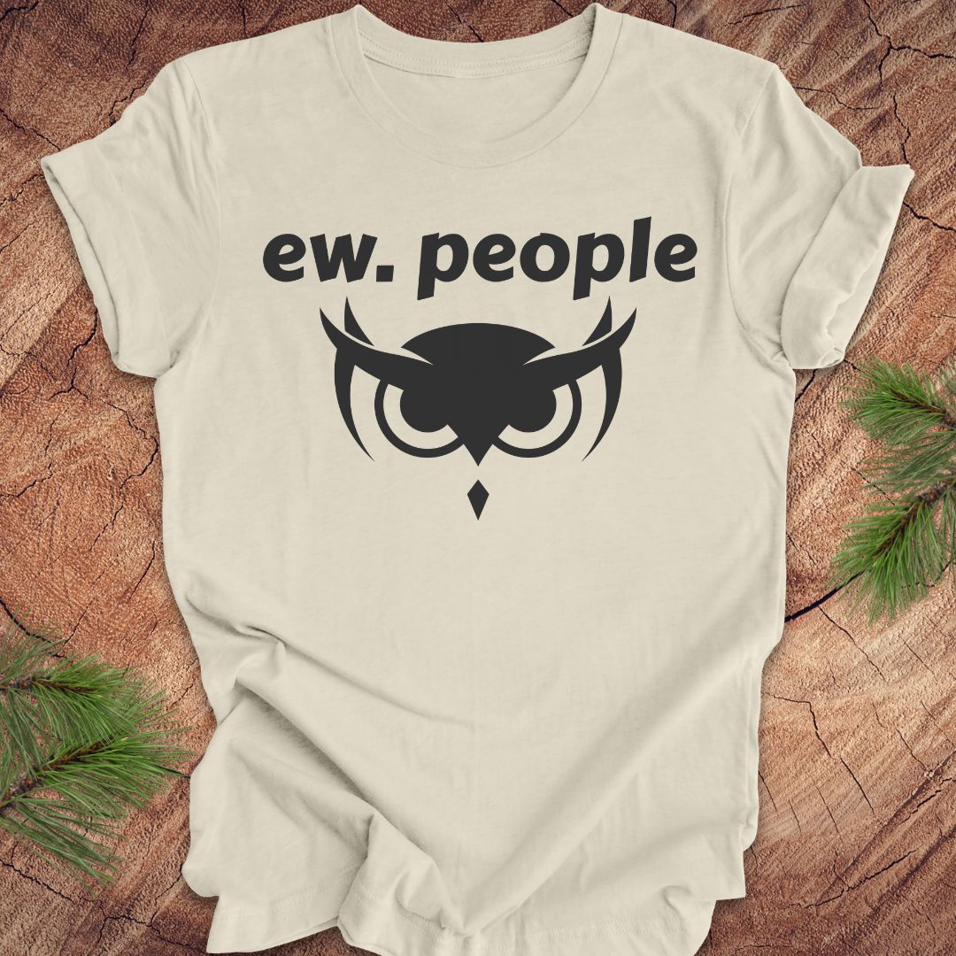 An off-white short-sleeve t-shirt with a black graphic print reading 'ew. people' above an owl face design, laid flat on a rustic wood background with pine branches in the corners.