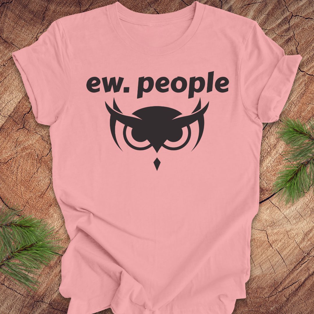 A light pink short-sleeve t-shirt with a black graphic print reading 'ew. people' above an owl face design, laid flat on a rustic wood background with pine branches in the corners.
