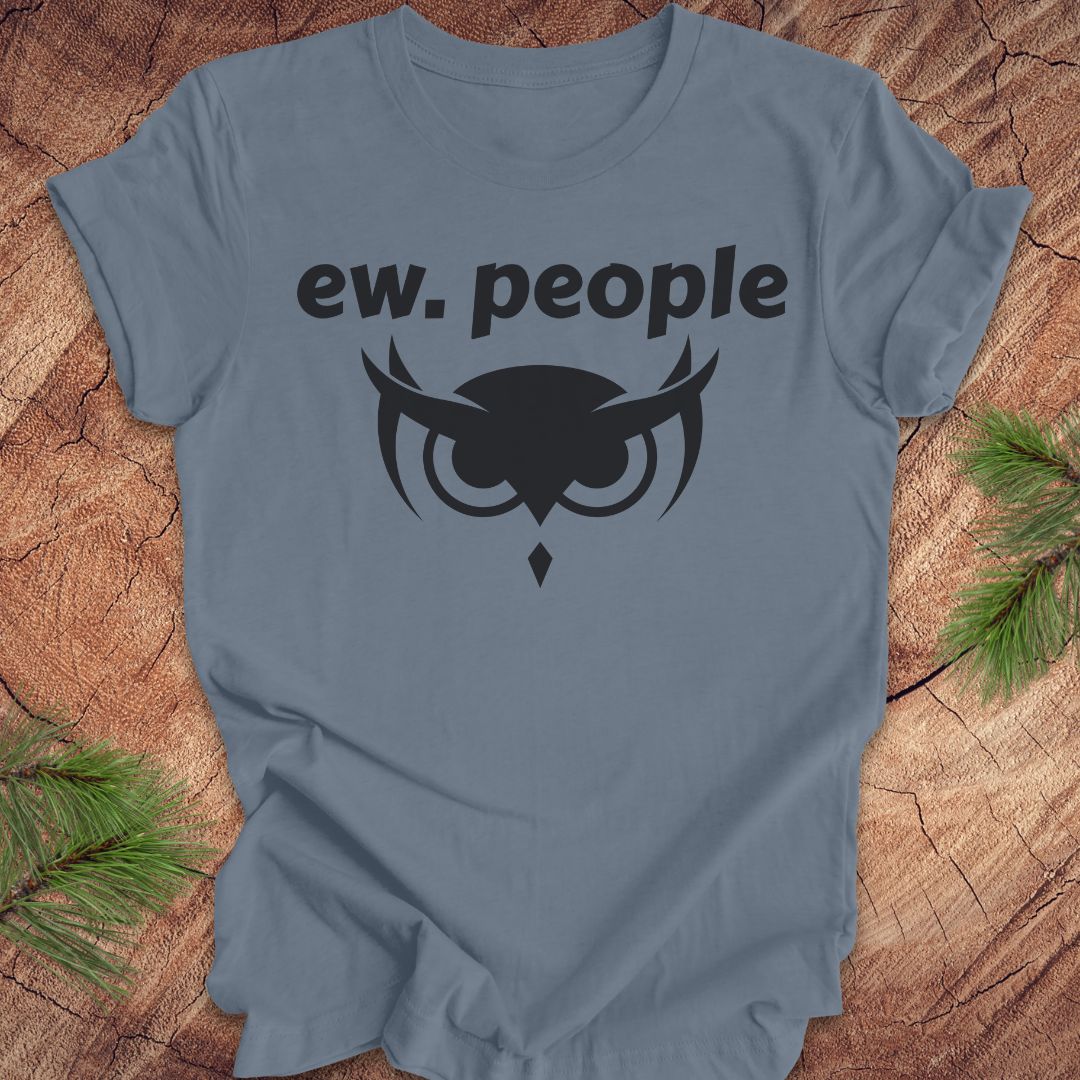 A slate blue crewneck t-shirt with a black graphic print that reads 'ew. people' above an owl design, laid flat on a rustic wood background with pine branches in the corners.