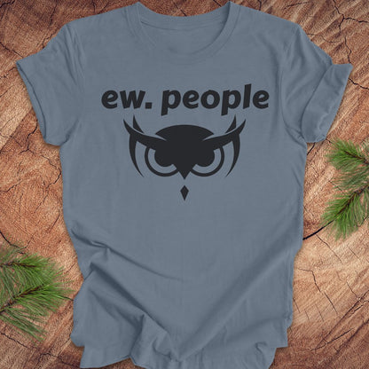 A slate blue crewneck t-shirt with a black graphic print that reads 'ew. people' above an owl design, laid flat on a rustic wood background with pine branches in the corners.