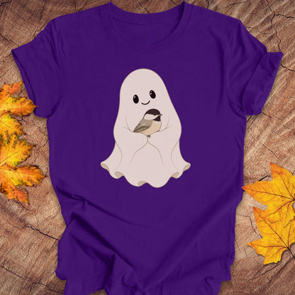 A purple short-sleeve t-shirt laid flat on a rustic wood surface, featuring a cute cartoon ghost graphic holding a small bird in the center. Yellow and orange autumn leaves are placed around the shirt as styling props.