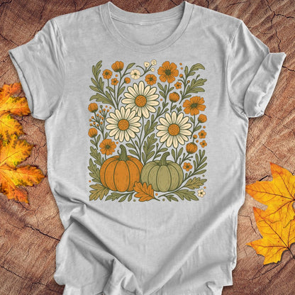 A light gray short-sleeve t-shirt with a retro autumn graphic of daisies, pumpkins, and fall leaves, laid flat on a rustic wood background with scattered yellow leaves.