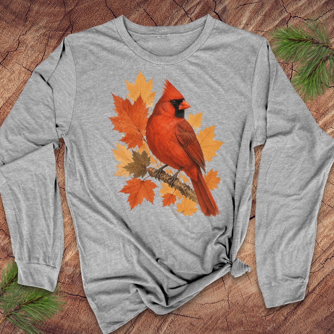 A gray long-sleeve t-shirt with a graphic of a red cardinal bird surrounded by orange and yellow autumn leaves, laid flat on a textured wood background with pine branches.