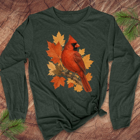 A dark forest green long-sleeve t-shirt with a graphic of a red cardinal bird surrounded by orange and yellow autumn leaves, laid flat on a textured wood background with pine branches.