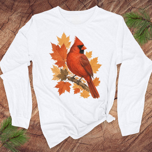 A white long-sleeve t-shirt with a graphic of a red cardinal bird surrounded by orange and yellow autumn leaves, laid flat on a textured wood background with pine branches.
