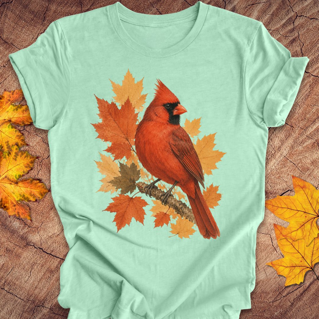 A mint green short-sleeve t-shirt laid flat on a rustic wood background, featuring a graphic of a red cardinal perched on orange and brown autumn maple leaves.