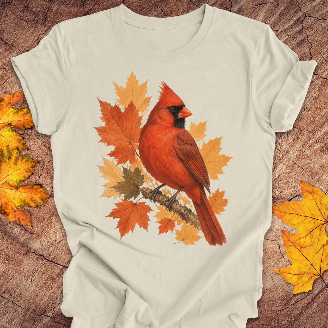 An beige short-sleeve t-shirt with a graphic of a red cardinal perched on autumn maple leaves, laid flat on a rustic wood background with scattered fall leaves.
