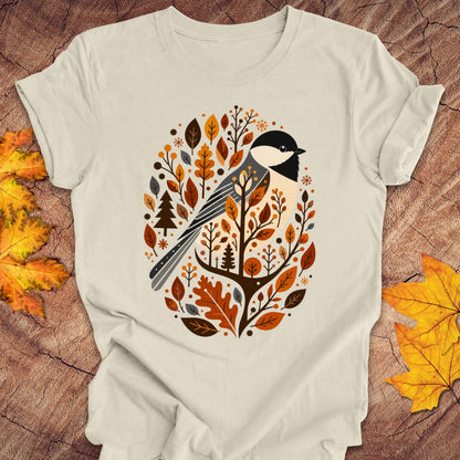 An off-white short-sleeve t-shirt with a graphic of a bird surrounded by autumn leaves, styled flat on a rustic wood background with yellow maple leaves.