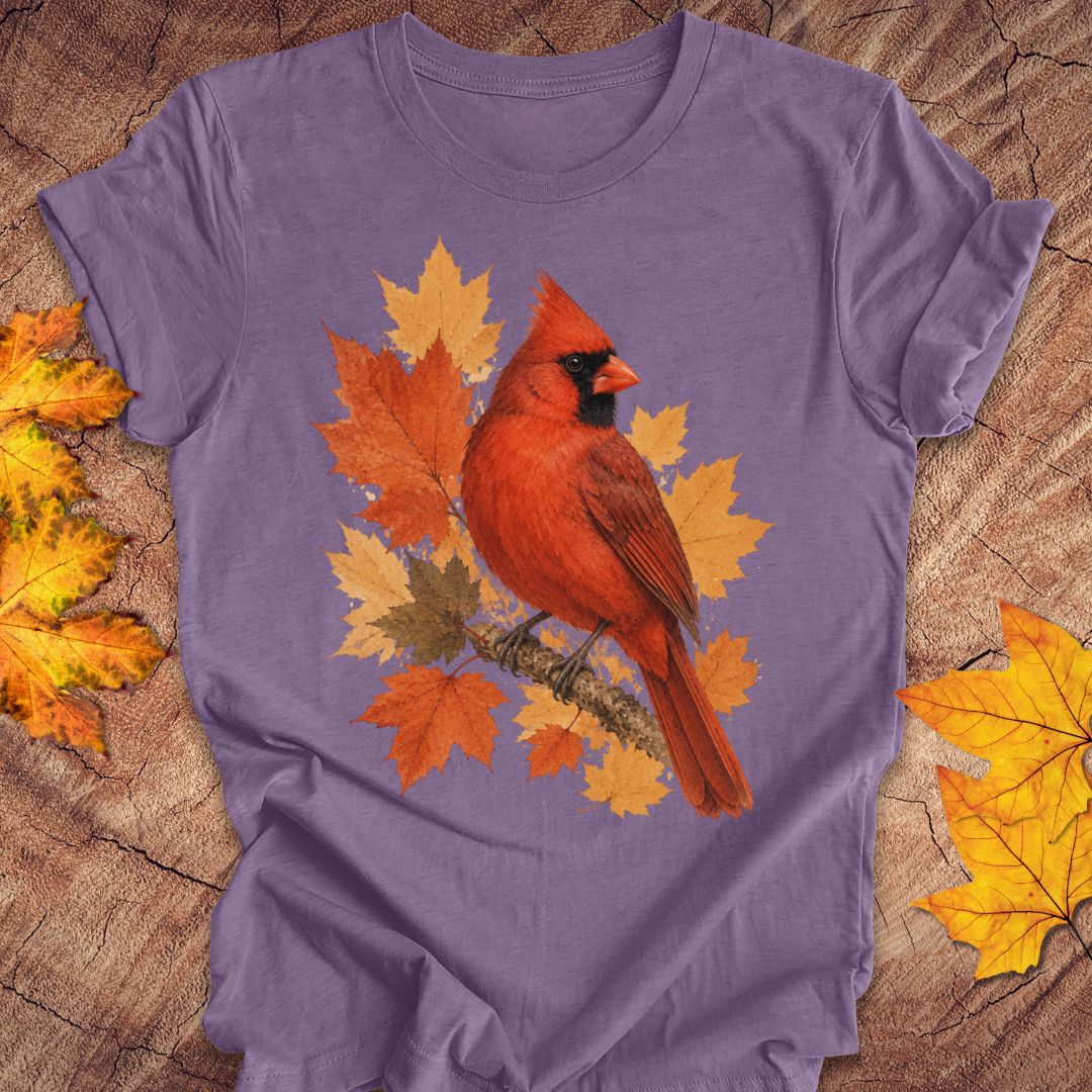 A purple short-sleeve t-shirt laid flat on a rustic wood surface, featuring a graphic of a red cardinal perched among orange and yellow autumn leaves. Decorative yellow maple leaves are placed in the corners of the frame.