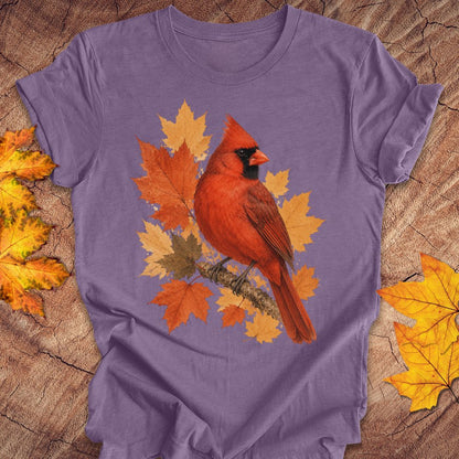 A purple short-sleeve t-shirt laid flat on a rustic wood surface, featuring a graphic of a red cardinal perched among orange and yellow autumn leaves. Decorative yellow maple leaves are placed in the corners of the frame.