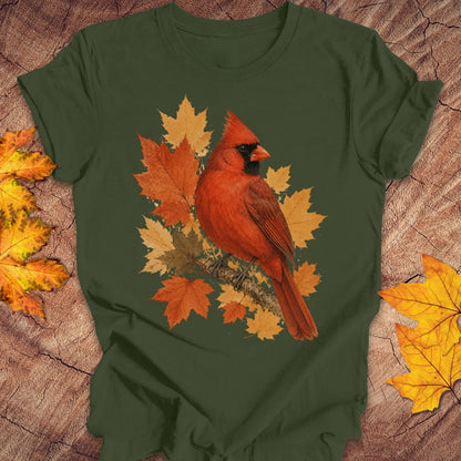 An olive green short-sleeve t-shirt with a graphic of a red cardinal perched on autumn maple leaves, laid flat on a rustic wood background with scattered yellow and orange leaves.