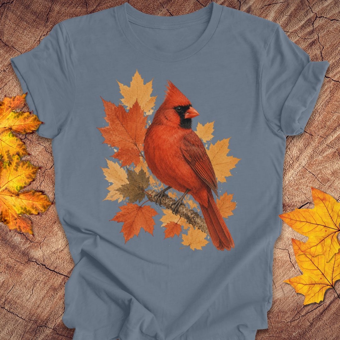 A slate blue crewneck t-shirt featuring a graphic print of a red cardinal perched on orange and yellow autumn maple leaves. The t-shirt is laid flat on a rustic wood background, framed by scattered yellow and orange leaves in the corners.