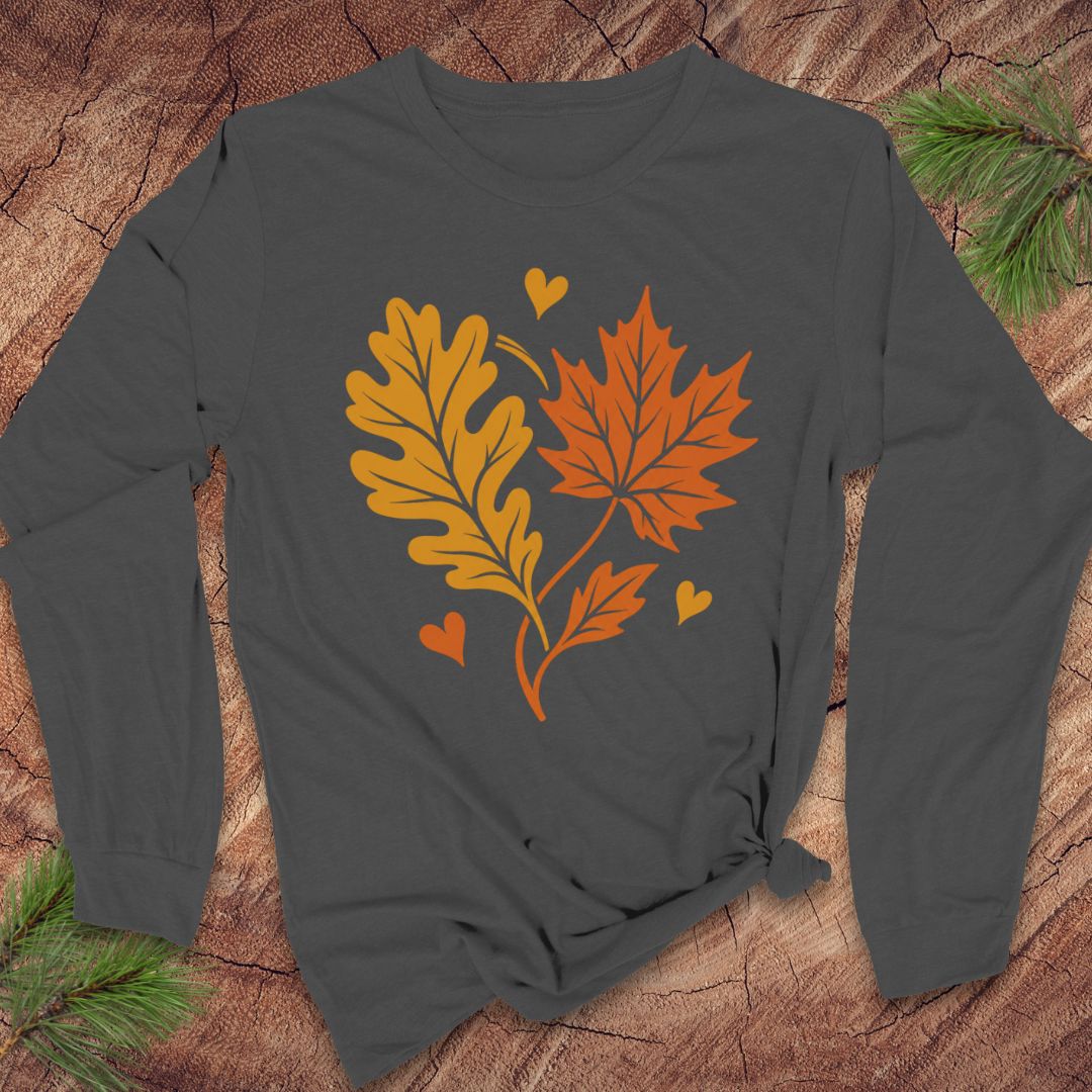 A dark charcoal gray long-sleeve t-shirt with a graphic of an orange maple leaf and a yellow-orange oak leaf with small hearts, laid flat on a rustic wood background with pine branches.