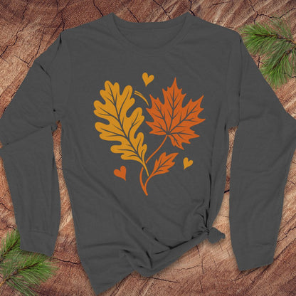 A dark charcoal gray long-sleeve t-shirt with a graphic of an orange maple leaf and a yellow-orange oak leaf with small hearts, laid flat on a rustic wood background with pine branches.