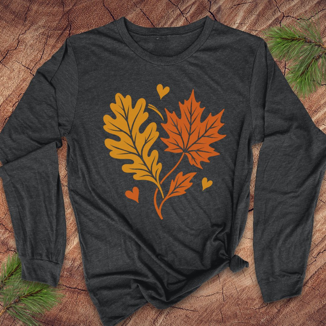 A dark charcoal gray long-sleeve t-shirt with a graphic of an orange maple leaf and a yellow-brown oak leaf with small orange and yellow hearts, laid flat on a rustic wood background with pine branches.