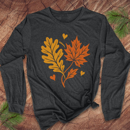 A dark charcoal gray long-sleeve t-shirt with a graphic of an orange maple leaf and a yellow-brown oak leaf with small orange and yellow hearts, laid flat on a rustic wood background with pine branches.
