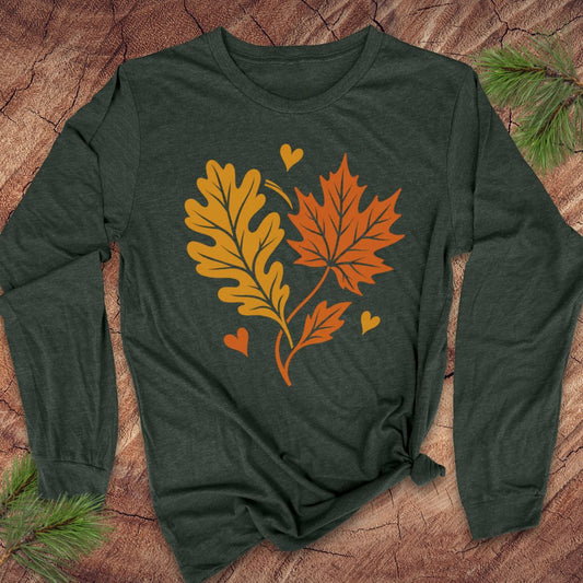 A dark forest green long-sleeve t-shirt with a graphic of autumn leaves (an oak leaf in yellow, a maple leaf in orange, and three small hearts in orange and yellow) on the front. The shirt is styled flat on a textured wood background with pine branches in the corners.