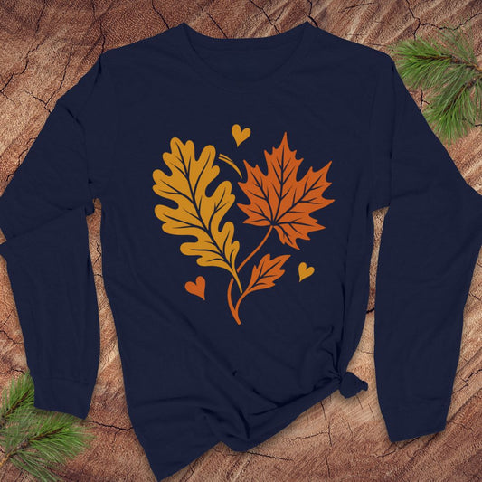 A navy blue long-sleeve t-shirt with a graphic of autumn leaves (an oak leaf and a maple leaf) and small hearts in orange and yellow, laid flat on a rustic wood background with pine needles.