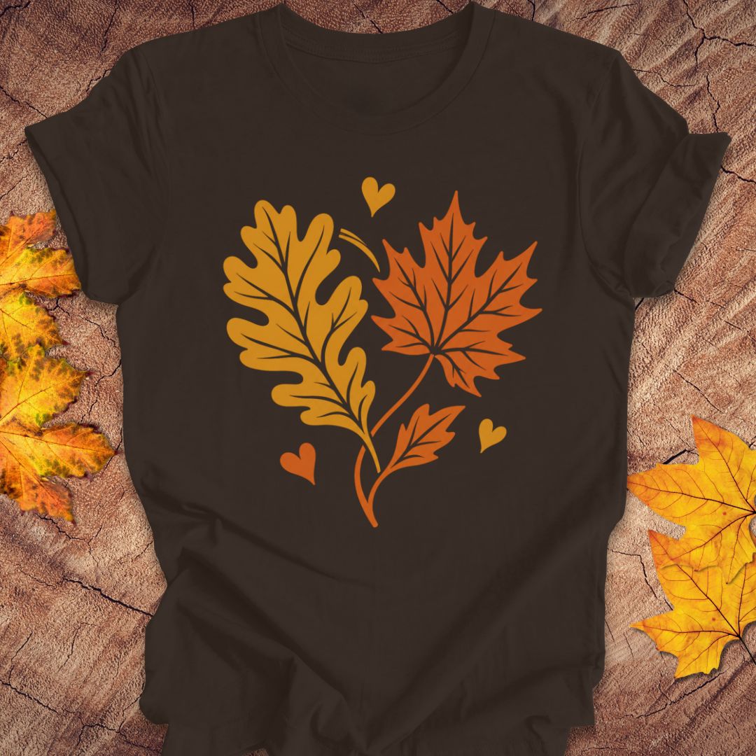 A brown short-sleeve t-shirt laid flat on a rustic wood surface, featuring a graphic of two autumn leaves and three small hearts in orange and yellow. Decorative yellow and orange leaves are placed around the edges of the frame.