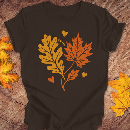 A brown short-sleeve t-shirt laid flat on a rustic wood surface, featuring a graphic of two autumn leaves and three small hearts in orange and yellow. Decorative yellow and orange leaves are placed around the edges of the frame.
