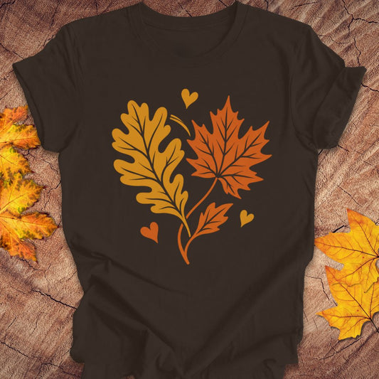 A brown short-sleeve t-shirt laid flat on a rustic wood surface, featuring a graphic of two autumn leaves and three small hearts in orange and yellow. Decorative yellow and orange leaves are placed around the edges of the frame.