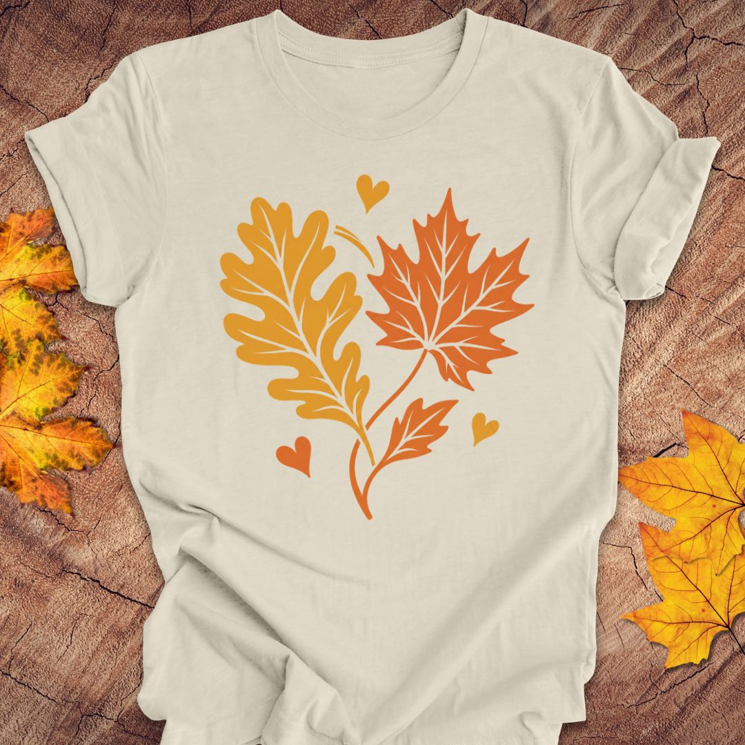 An beige short-sleeve t-shirt with a graphic of two autumn leaves (an oak leaf in yellow-orange and a maple leaf in orange) with small hearts, laid flat on a rustic wood background with decorative fall leaves.
