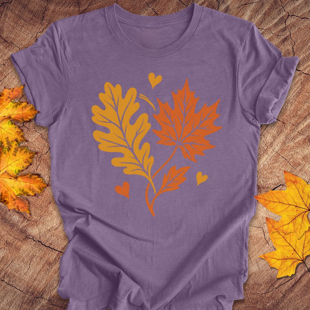 A purple short-sleeve t-shirt laid flat on a rustic wood surface, featuring a graphic of two orange autumn leaves and three small hearts. Yellow-orange leaves are also scattered around the edges of the frame as decorative props.