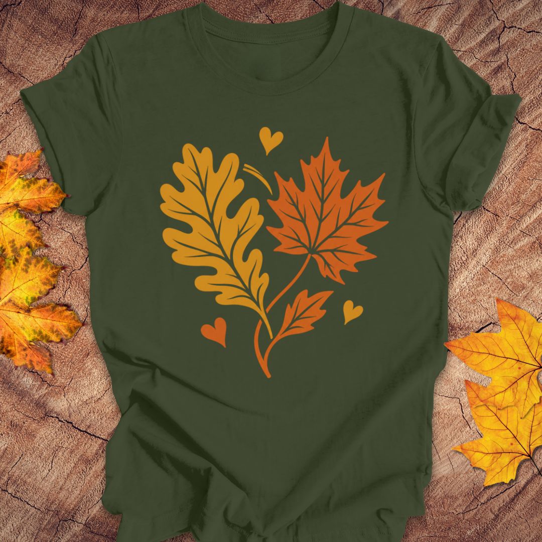 An olive green short-sleeve t-shirt laid flat on a rustic wood background, featuring a graphic of two autumn leaves (an oak leaf in mustard yellow and a maple leaf in orange) with small hearts.