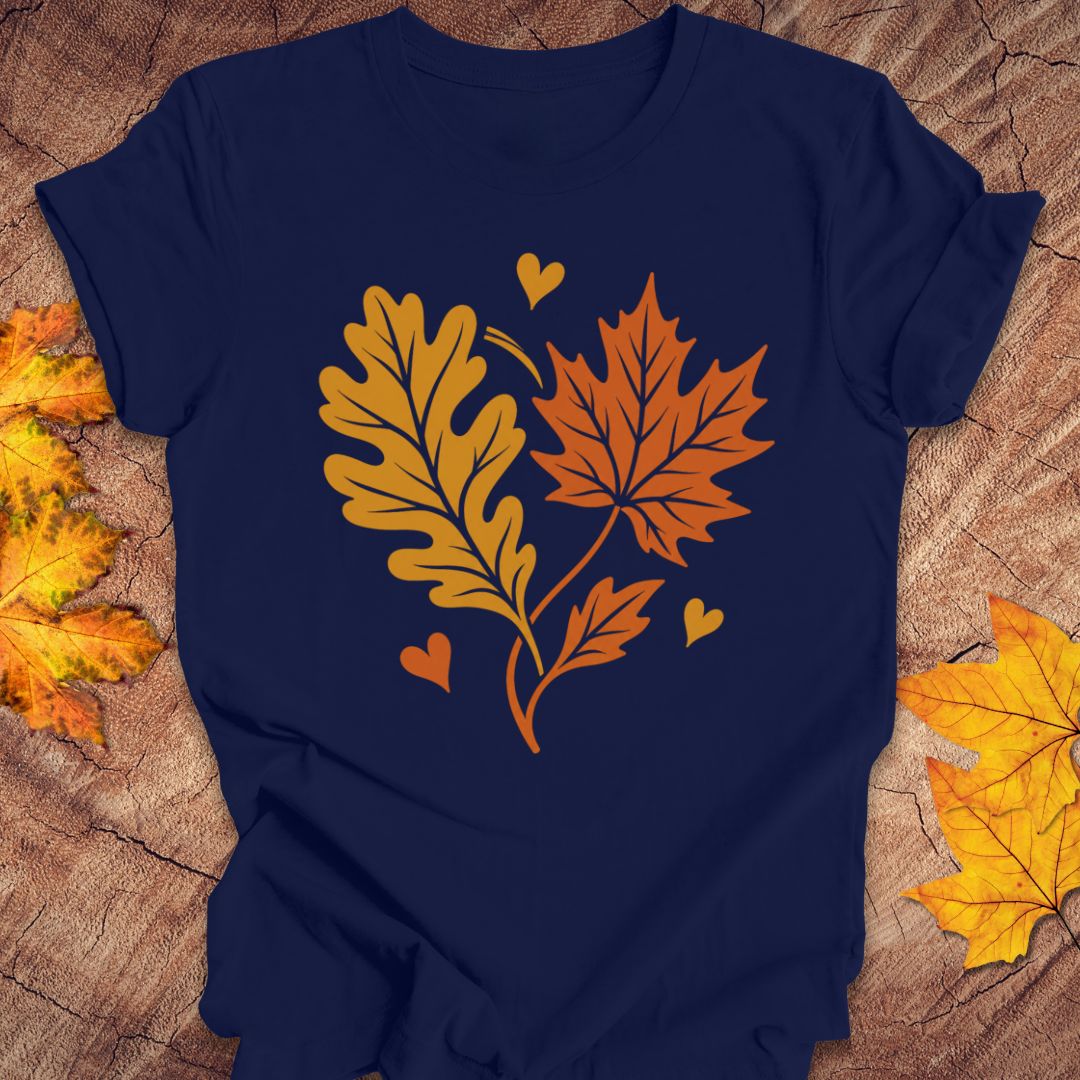 A navy blue short-sleeve t-shirt laid flat on a rustic wood surface, featuring a graphic of two orange autumn leaves and three small hearts. Yellowed decorative leaves are placed around the edges of the frame.
