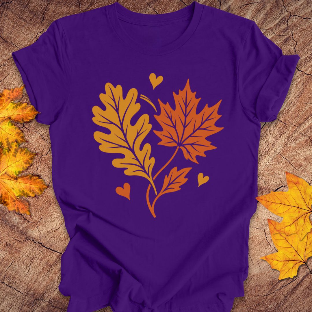 A purple short-sleeve t-shirt laid flat on a rustic wood surface, featuring a graphic of an orange maple leaf and a yellow-orange oak leaf with small hearts on the front. Yellow and orange autumn leaves are placed around the edges of the frame.