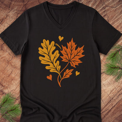 A black V-neck t-shirt laid flat on a rustic wood background with pine branches in the corners. The shirt features a graphic of two autumn leaves, one orange-brown oak leaf and one red-orange maple leaf, forming a heart shape with small yellow hearts.