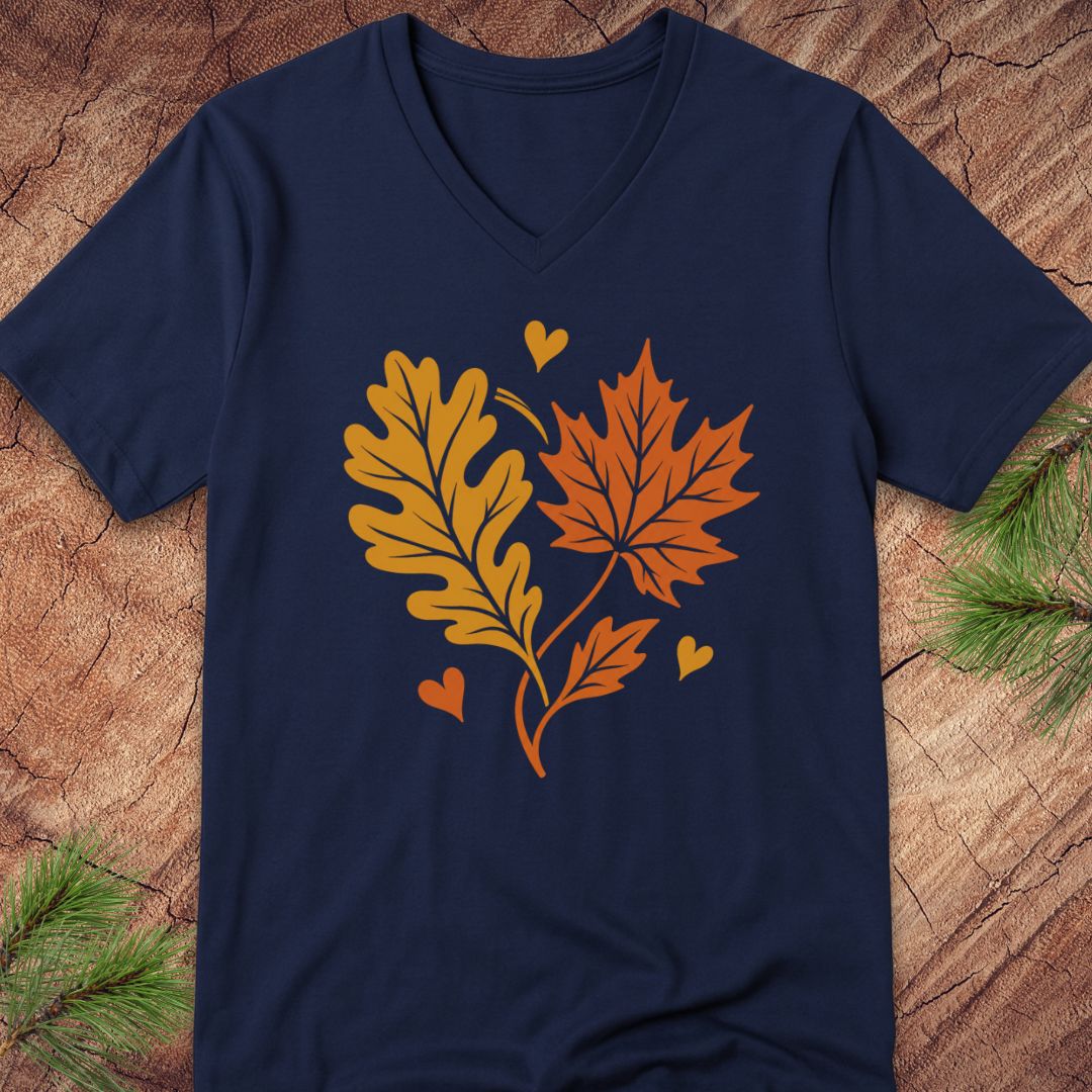 A navy blue V-neck t-shirt featuring a graphic of autumn leaves (an oak leaf in yellow and a maple leaf in orange) with small hearts, laid flat on a rustic wood background with pine branches.