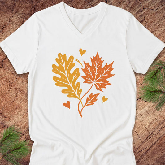 A white V-neck t-shirt laid flat on a rustic wood background with pine branches in the corners. The shirt features a graphic of an orange maple leaf and a yellow-orange oak leaf with small orange and yellow hearts.