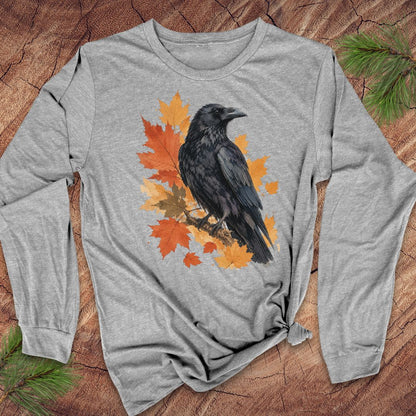 A heather gray long-sleeve t-shirt with a graphic of a black raven perched on orange and yellow autumn maple leaves, laid flat on a rustic wood background with pine needles.