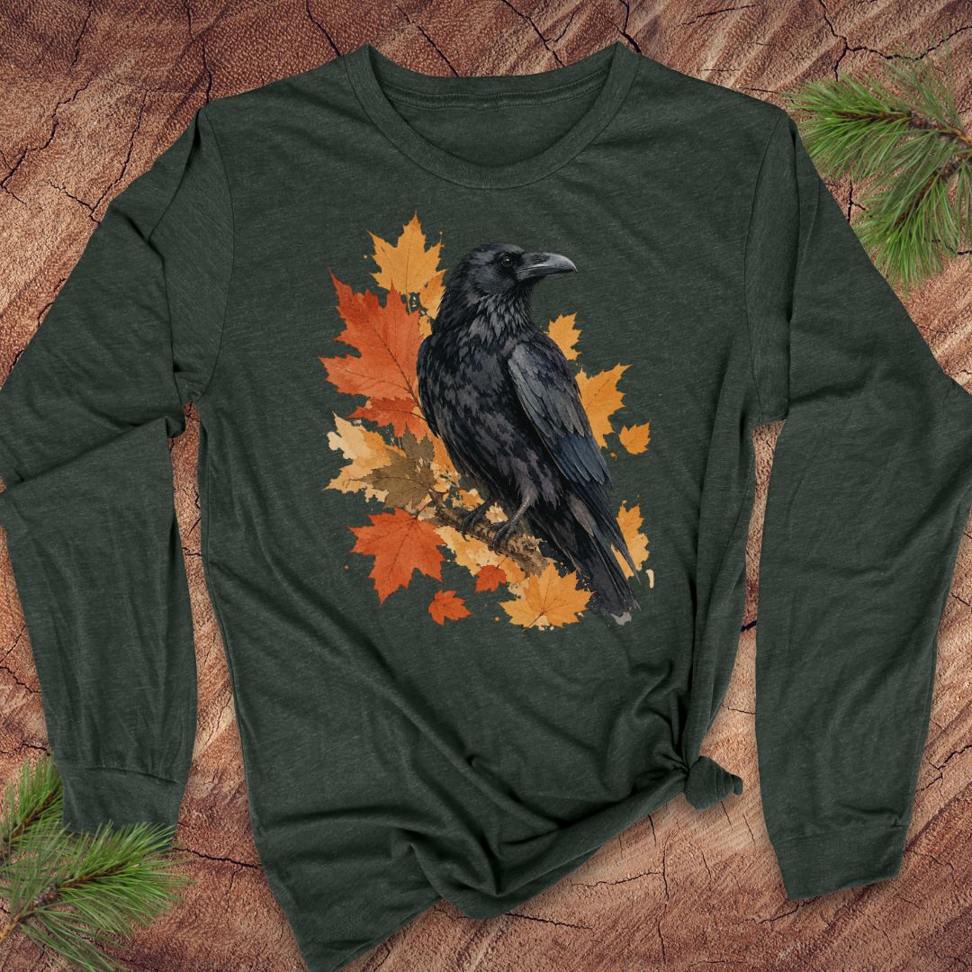 A dark forest green long-sleeve t-shirt with a graphic of a black raven perched on orange and yellow autumn leaves, laid flat on a rustic wood background with pine branches.