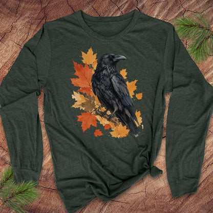 A dark forest green long-sleeve t-shirt with a graphic of a black raven perched on orange and yellow autumn leaves, laid flat on a rustic wood background with pine branches.