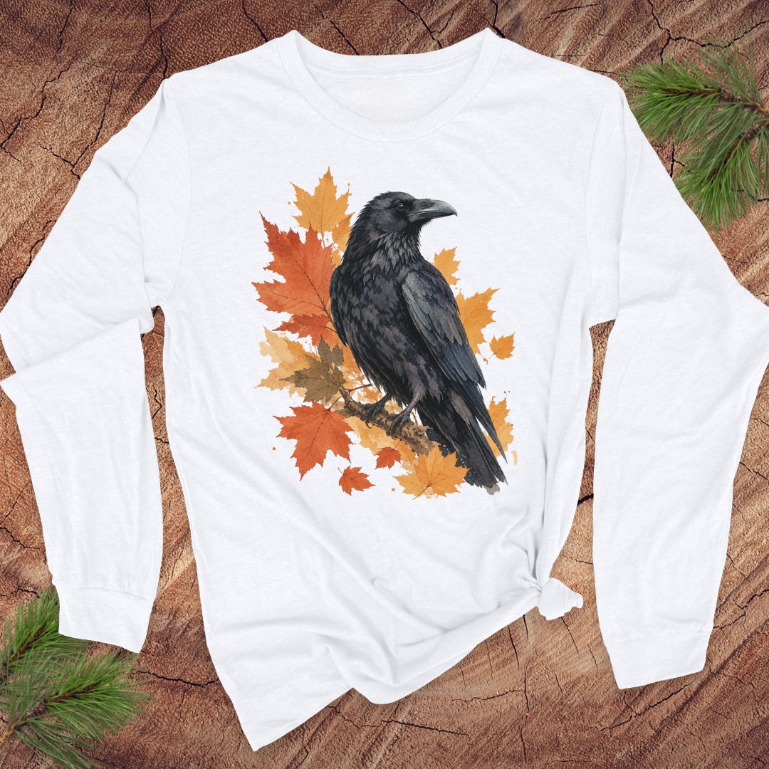 A white long-sleeve t-shirt with a graphic of a black raven perched on orange and yellow autumn leaves, laid flat on a rustic wood background with pine branches.
