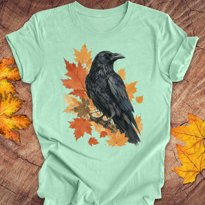 A mint green short-sleeve t-shirt with a graphic of a black raven perched on orange and yellow autumn leaves, laid flat on a rustic wood background with scattered fall leaves.