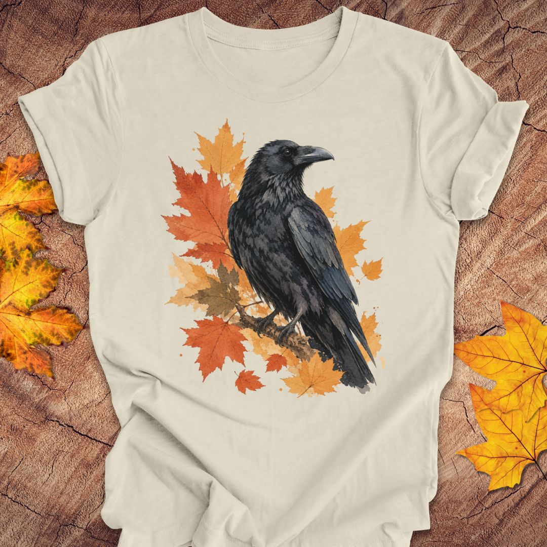 A beige short-sleeve t-shirt with a graphic print of a black raven perched on orange and yellow autumn maple leaves, laid flat on a rustic wood background with scattered fall leaves.