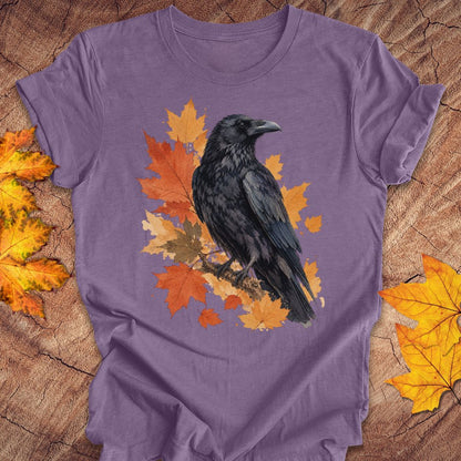 A purple short-sleeve t-shirt laid flat on a rustic wood surface, featuring a graphic of a black raven perched on orange and yellow autumn leaves. Decorative yellow maple leaves are placed in the corners of the wood background.
