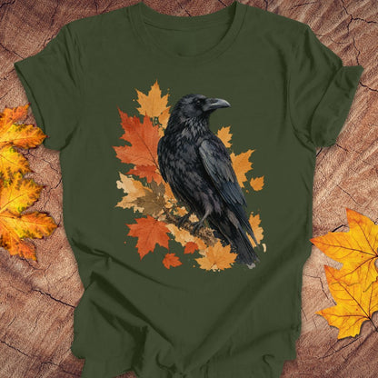 An olive green short-sleeve t-shirt with a graphic of a black raven perched on orange and yellow autumn leaves, laid flat on a rustic wood background with scattered fall leaves.