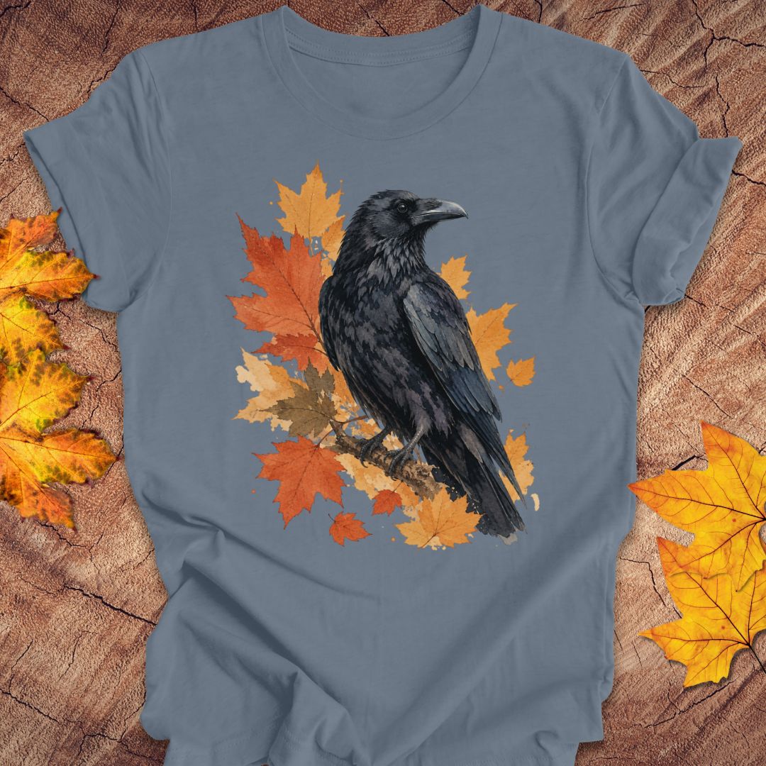 A slate blue crewneck t-shirt featuring a graphic of a black raven perched on orange and yellow autumn leaves, laid flat on a rustic wood background with scattered fall leaves.