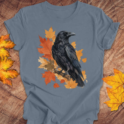 A slate blue crewneck t-shirt featuring a graphic of a black raven perched on orange and yellow autumn leaves, laid flat on a rustic wood background with scattered fall leaves.