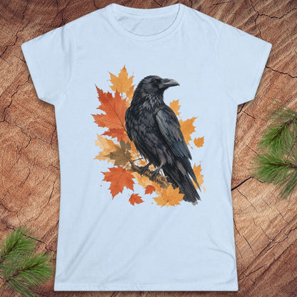 A light blue short-sleeve crewneck t-shirt featuring a graphic print of a black raven perched on orange and yellow autumn maple leaves, displayed flat on a rustic wood background with pine branches in the corners.