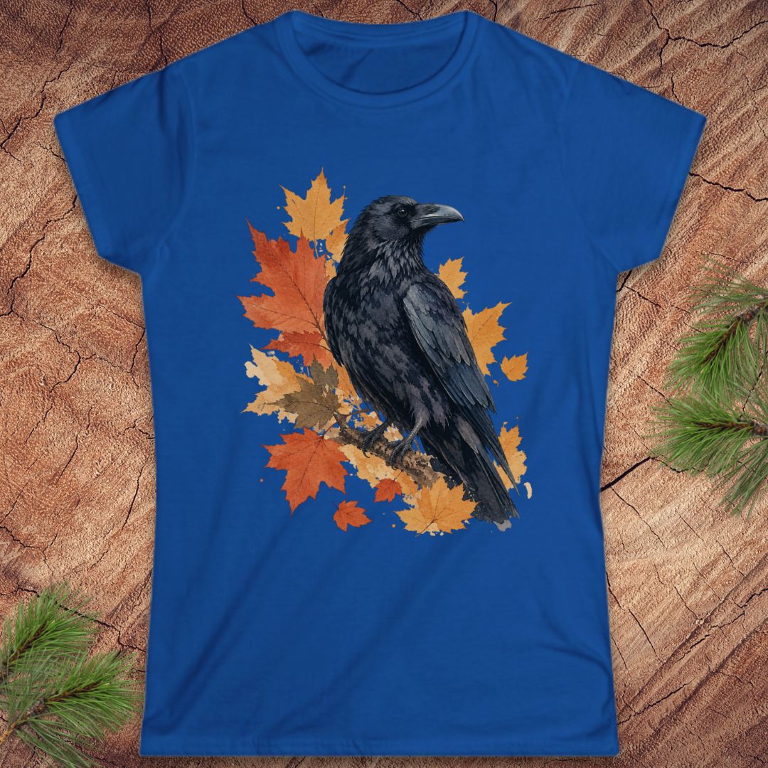 A royal blue short-sleeve t-shirt with a graphic of a black raven perched on orange and yellow autumn maple leaves, laid flat on a textured wood background with pine branches in the corners.