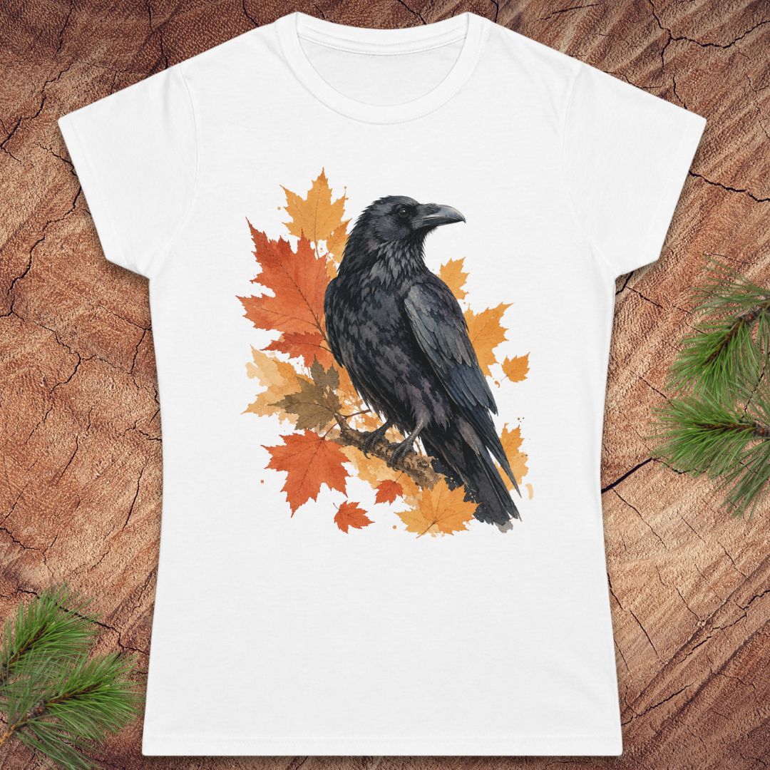 A white short-sleeve crewneck t-shirt featuring a graphic print of a black raven perched on orange and yellow autumn maple leaves, displayed flat on a rustic wood background with pine branches in the corners.