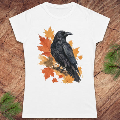A white short-sleeve crewneck t-shirt featuring a graphic print of a black raven perched on orange and yellow autumn maple leaves, displayed flat on a rustic wood background with pine branches in the corners.