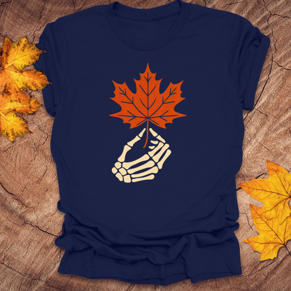 Navy blue t-shirt with a skeleton hand holding an orange maple leaf on a wooden surface with fall leaves.