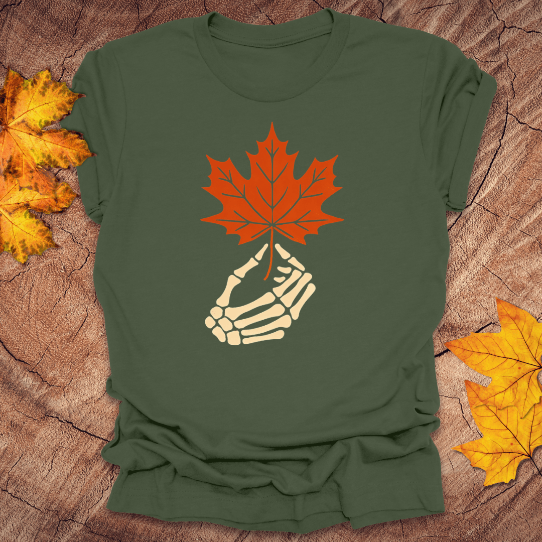 Green t-shirt with a skeleton hand holding an orange maple leaf on a wooden surface with fall leaves.